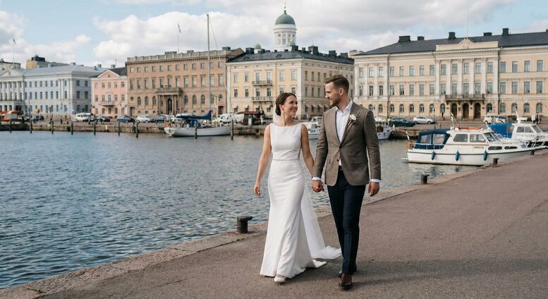 A bride and groom holding hands on the steps of the iconic Helsinki Cathedral at Senate Square during their sunny city wedding, with a vintage car parked below