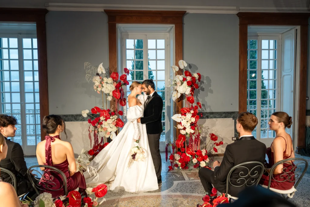 Indoor wedding ceremony inside the grand hall of Villa Cipressi, featuring high ceilings, floor-to-ceiling windows with lake views, and red floral arrangements.