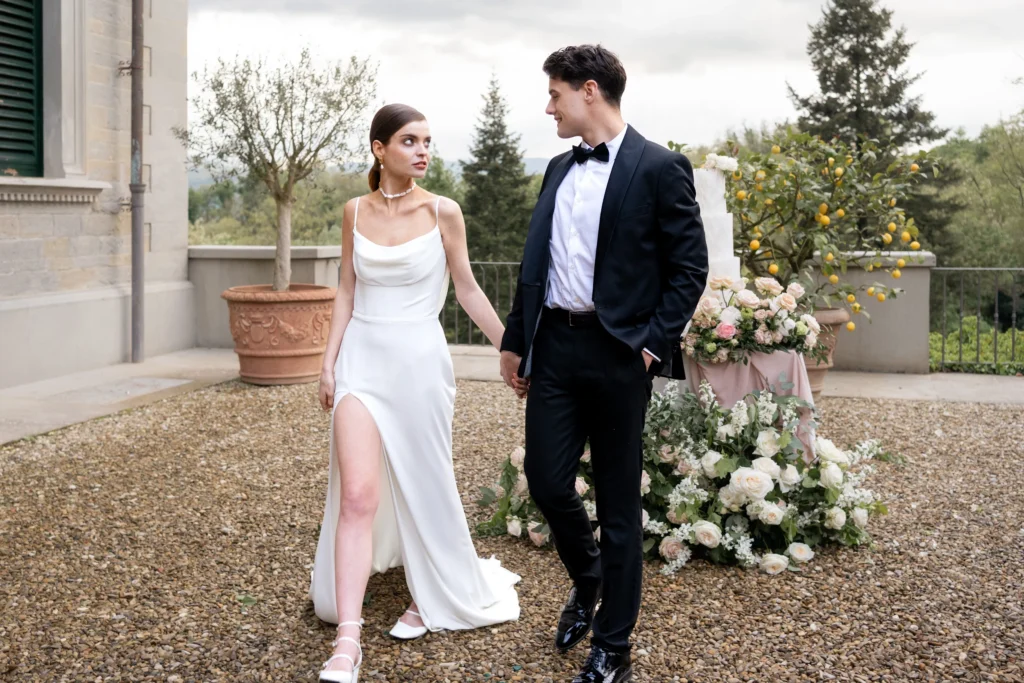 A bride and groom walking hand-in-hand on a gravel terrace next to a large tiered wedding cake and potted lemon trees, with the villa wall on the left. 