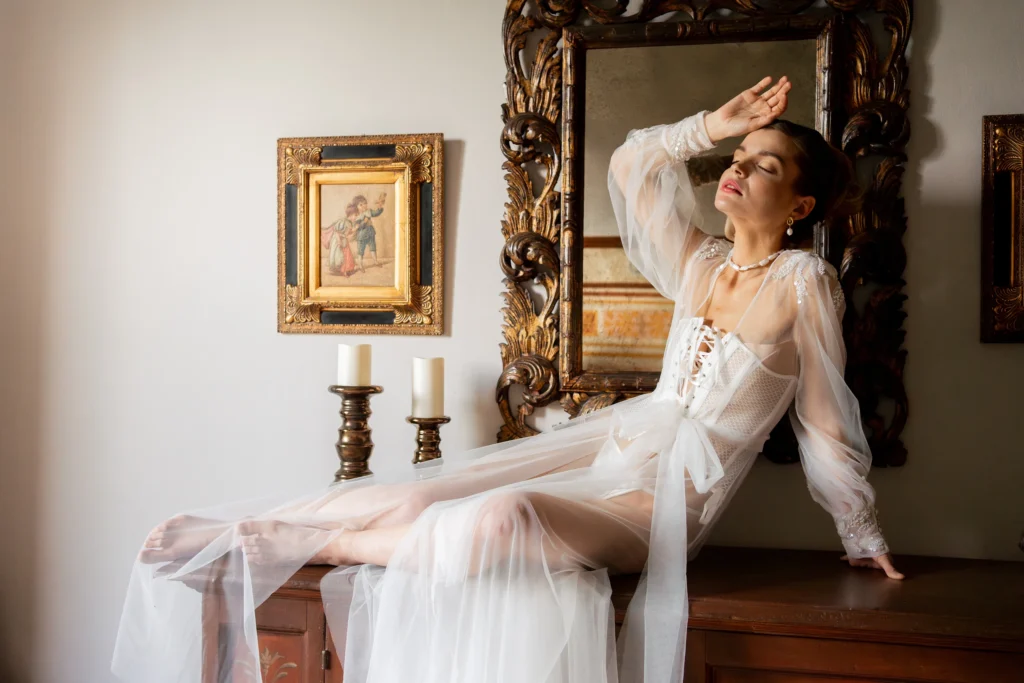 A bride wearing a sheer white dressing gown sitting on a wooden antique chest of drawers in a sunlit room with a mirror and candles, capturing a quiet moment before the ceremony. 