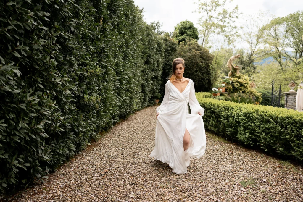 A bride in a long white dress walking joyfully along a gravel path lined with tall green hedges, holding her dress up slightly. 