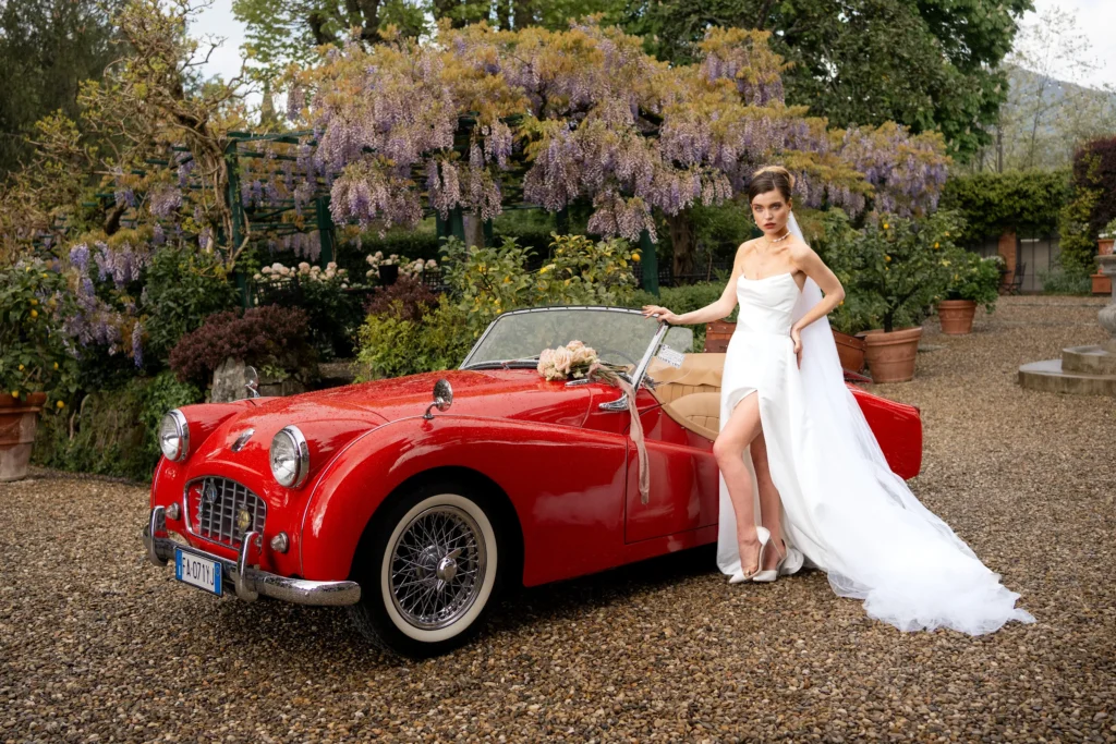 A bride in a modern white wedding dress posing elegantly by leaning against a red vintage convertible car, with wisteria flowers and villa gardens in the background